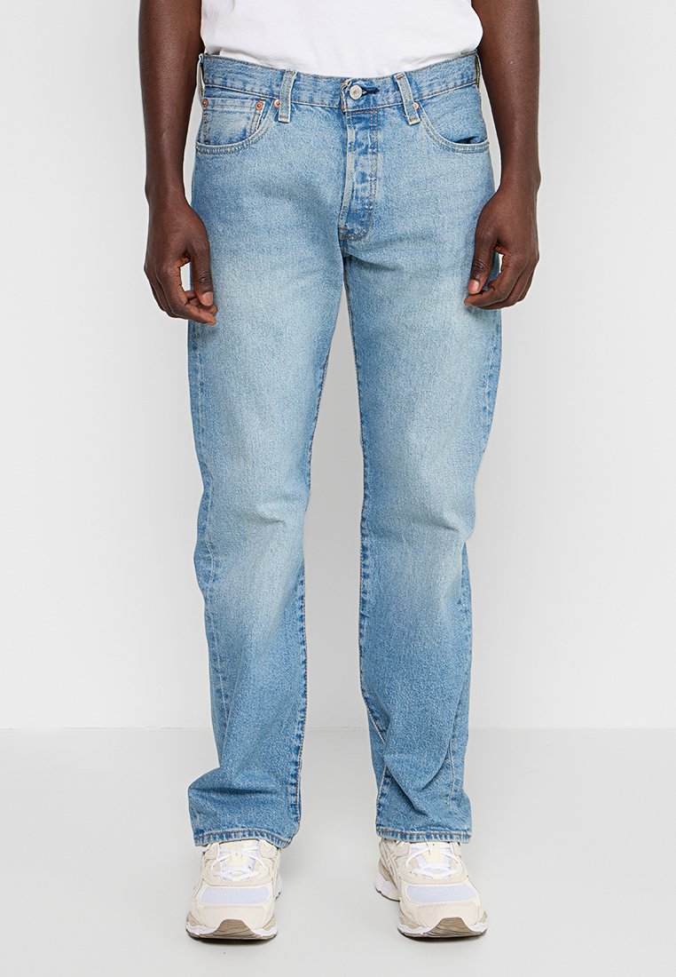 Helle blaue Jeans mit geradem Schnitt, verblasster Optik, Fünf-Taschen-Design und Knopfverschluss aus Metall. Getragen mit weißen Turnschuhen.