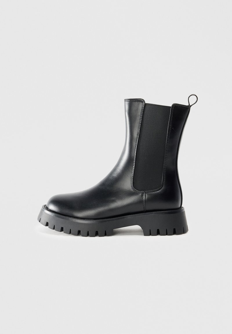 Botte Chelsea en cuir noir avec semelle crantée épaisse, panneau latéral élastique et languette de tirage sur fond gris.