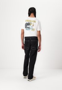 White t-shirt with colorful graphic print on back, paired with black pants featuring white stitching and two back pockets. Neutral sneakers.