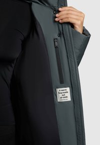 Podložena jakna iz temnosive tkanine, ki ima žep z zadrgo in oznako z napisom "Topel objem iz blaga" znotraj.