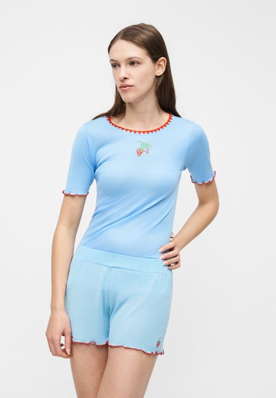 Γυναίκα που φοράει ανοιχτό μπλε τοπ με κοντά μανίκια και ταιριαστά σορτς με κόκκινα κυματοειδή τελειώματα και κεντήματα φράουλας.
