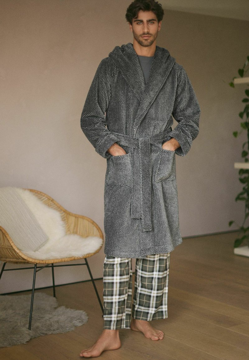 Grå, tekstureret badekåbe med hætte og snoreliv, der har to frontlommer; båret over rutede pyjamasbukser i grøn og hvid.
