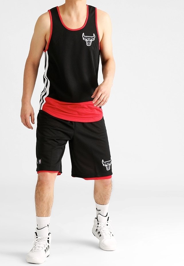 Homme portant un maillot et un short de basket-ball Chicago Bulls noir et rouge, des chaussettes blanches et des chaussures de sport blanches, debout devant un fond uni.
