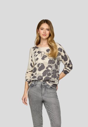 Frau mit langem blondem Haar, die einen beige-grau gepunkteten Pullover mit Strassbuchstaben und graue, hoch taillierte Jeans vor grauem Hintergrund trägt.