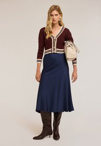 Cardigan lavorato a maglia bordeaux con strisce bianche, chiusura a bottoni, abbinato a una gonna midi di satin blu navy e stivali marroni al ginocchio. Borsa beige.