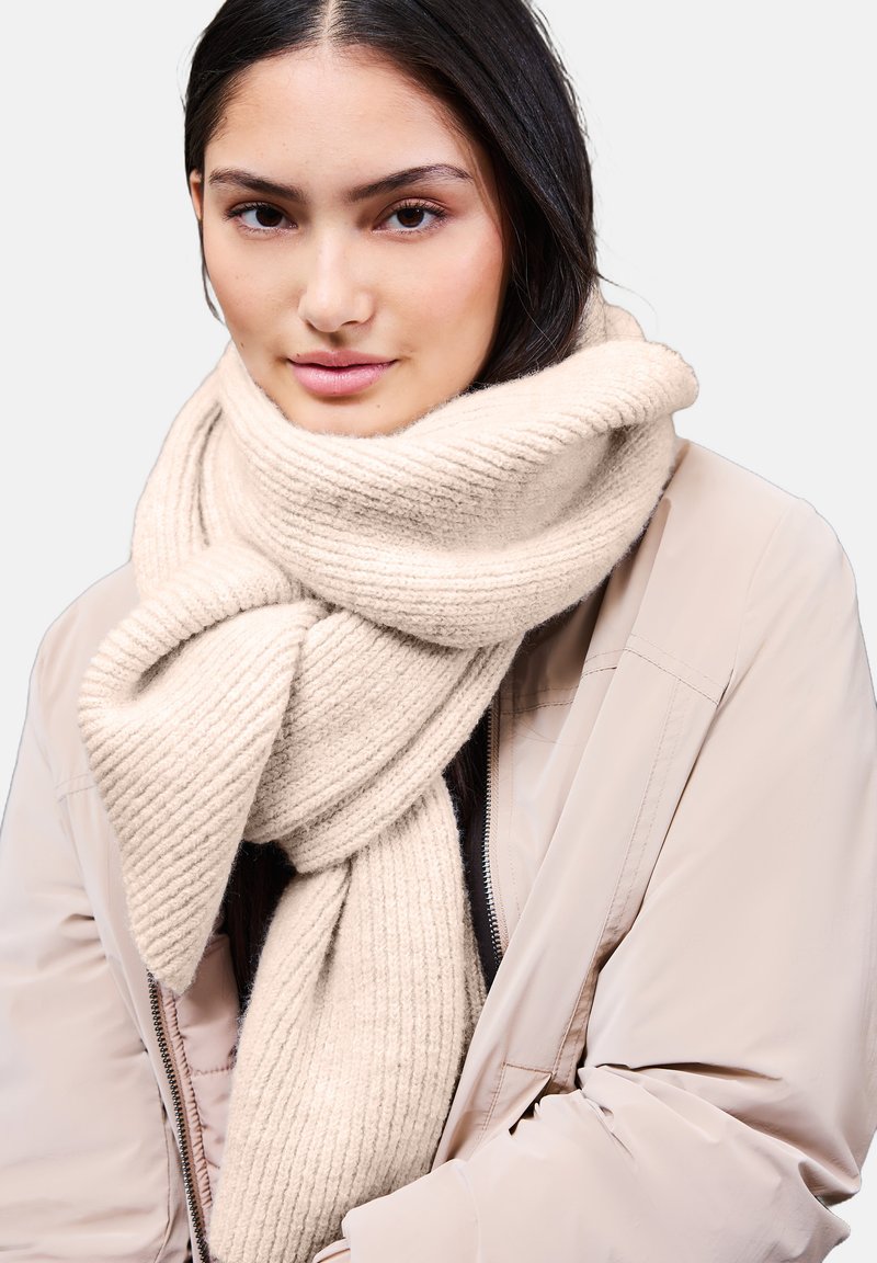 Bufanda chunky de punto acanalado en suave tejido beige, enrollada de manera suelta alrededor del cuello, combinada con una chaqueta bomber de color claro.
