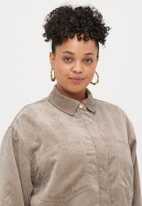 Šviesiai rudas kordūrovo marškinis su apykakle, dviem priekinių kišenėmis ir vario mygtukų detalėmis. Minkšta tekstūra su vertikaliu briaunojimu.