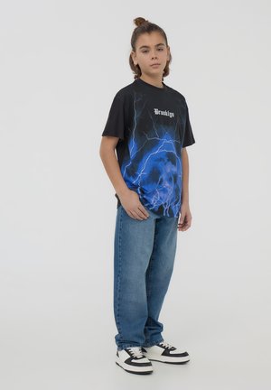 T-shirt grafica nera e blu con design di fulmini e testo "Brooklyn", abbinata a jeans blu e sneakers bianche e nere.