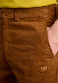 Ruskeat kordurohousut teksturoidulla pinnalla, joissa on etutasku ja vasemmalla puolella pieni kirjailtu Nike-logo.
