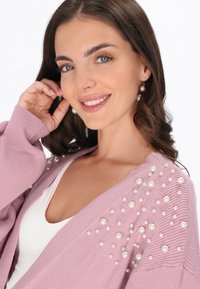 Ljusrosa kofta med ribbad textur och spridda konstpärlor på ena axeln. Rund halsringning med en vit top underneath.