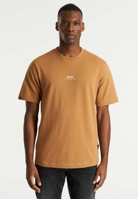 T-shirt marrone a maniche corte con colletto tondo, caratterizzato da un logo bianco sul petto. Tessuto testurizzato con vestibilità rilassata. Indossato con jeans scuri.