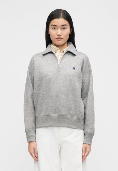 Sivi pulover iz flisa z zadrgo ovratnikom, rebrastimi manšetami in spodnjim robom. Na levi strani prsnega koša je majhen moder vezen logotip. Pod njim so oblečene bele hlače.