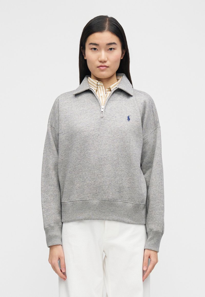 Pull-over en polaire grise avec col zippé, poignets et ourlet côtelés. Présente un petit logo brodé bleu sur le côté gauche de la poitrine. Pantalon blanc porté en dessous.