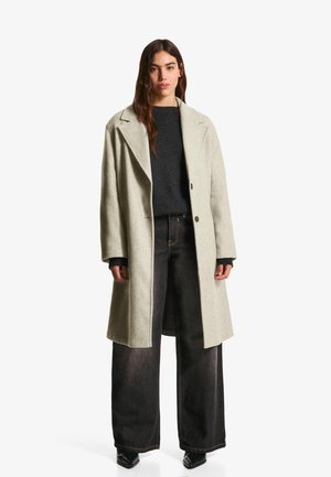 Frau trägt einen hellgrauen langen Mantel, einen schwarzen Pullover, weite schwarze Jeans und schwarze spitze Schuhe und steht vor einem schlichten weißen Hintergrund.