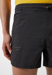 Schwarze Badebermuda aus leichtem Stoff, mit elastischem Bund, seitlicher Tasche und dezentem Branding auf der unteren linken Seite.