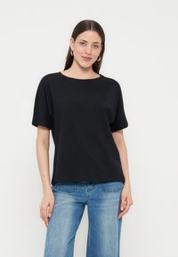 Γυναίκα με μακριά σκούρα μαλλιά φοράει χρυσούς κρίκους σκουλαρίκια, ένα φαρδύ μαύρο κοντομάνικο τοπ και ανοιχτό μπλε τζιν, στέκεται μπροστά σε λευκό φόντο.