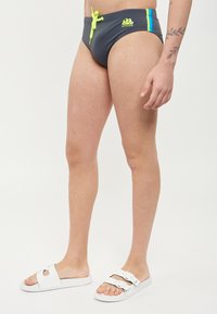 Grijze zwemslips met een neongele trekkoord, voorzien van blauwe en groene strepen aan de zijkanten. Gedragen met witte instap sandalen.