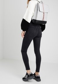 Harmaa reppu, jossa on läppä, vaaleanpunainen hihna ja musta pohja, käytettävissä mustien kireiden farkkujen ja mustien urheilukenkien kanssa, yksinkertaista taustaa vasten.