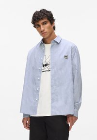 Modrá a biela kockovaná košeľa na gombíky, vyrobená z bavlny, voľný strih, s malou nášivkou loga na hrudi, nosená cez biele tričko s grafikou.