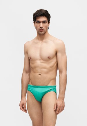 Mężczyzna o ciemnych włosach, ubrany w turkusowe kąpielówki, stoi na tle jednolitego jasnego tła, zwrócony twarzą do przodu, z neutralnym wyrazem twarzy.