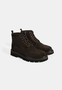 Bruine suède enkelboots met een ronde neus, vetersluiting aan de voorkant met metalen oogjes en een dikke zwarte rubberen zool. Geweven zijkanten en een lus om aan te trekken.