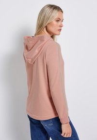 Sudadera de color rosa claro con un bolsillo frontal y capucha con cordón. Presenta tres rayas verticales grises a lo largo de las mangas. Material de mezcla de algodón.