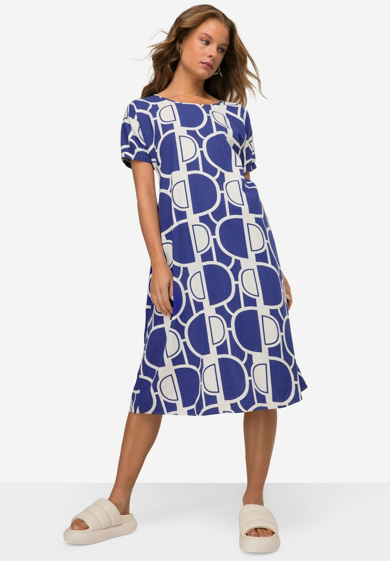 Modrá a bílá geometricky vzorovaná šaty s krátkým rukávem a uvolněným střihem, vyrobené z lehkého materiálu, v kombinaci s krémovými skluznými sandály.