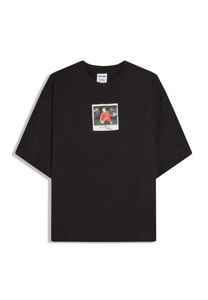 T-shirt nera oversize con una piccola grafica quadrata sul petto che raffigura un uomo con una giacca rossa accucciato tra due ruote di auto.