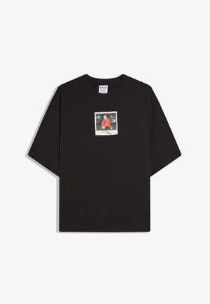 Fekete, bő fazonú póló, melynek mellkasán egy kis négyzet alakú grafikán egy piros dzsekis férfi guggol két autógumi között.