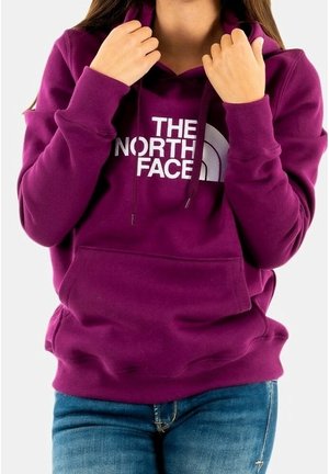 Persoon die de capuchon van een paarse The North Face-hoodie aanpast, gekleed in een blauwe spijkerbroek, voor een effen achtergrond.