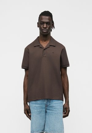 Άνδρας με σκουρόχρωμο δέρμα και κοντά dreadlocks, που φοράει καφέ κοντομάνικο πόλο και ανοιχτόχρωμα μπλε τζιν σορτς, στέκεται μπροστά σε απλό φόντο.