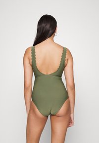 Fato de banho de uma peça verde-azeitona com alças em forma de escalope, design de costas baixas e uma textura suave. Minimalista, com uma silhueta ajustada.