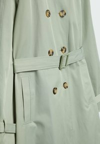 Trench-coat croisé vert clair avec des boutons marron, une ceinture à la taille avec boucle et des poches fendues verticales de chaque côté.