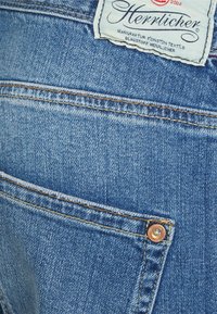 Jeans de denim azul claro com um bolso traseiro com um rebite de cobre e costura em contraste. A etiqueta diz "Herrlicher" com um acabamento texturizado.