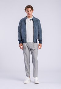 Chaqueta bomber de ante azul con cuello alto, camiseta gris de manga larga, pantalones grises y zapatillas blancas, exhibidos sobre un fondo sencillo.