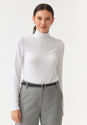 Weißes, geripptes Rollkragenoberteil mit langen Ärmeln, kombiniert mit grauen Hosen und einem schwarzen Gürtel mit einer kreisförmigen silbernen Schnalle. Schlichtes Design.