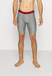 Graue gemusterte Boxershorts mit rotem Bund, der mit "UNDER ARMOUR" gekennzeichnet ist, im taillierten Design und bis zur Mitte des Oberschenkels.