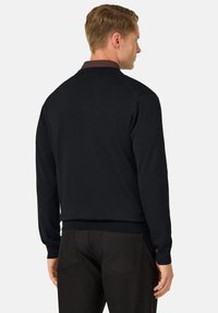 Pull en tricot noir à manches longues et col classique, présentant une texture lisse et une coupe ajustée. Porté par-dessus un pantalon sombre.