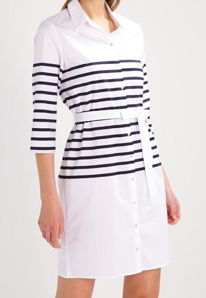 Kobieta ubrana w białą sukienkę koszulową w czarne poziome paski, z rękawami za łokieć, guzikami z przodu i paskiem wiązanym w talii.