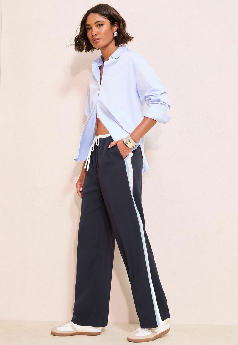 Žena pózuje v čiernych širokých nohaviciach s bielymi bočnými pruhmi, svetlo modrej oversized košeli s gombíkmi, béžových a bielych teniskách a minimalistickými šperkami.