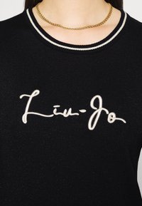 Zwarte geribbelde trui met een wit geborduurd "Liu-Jo" logo. Heeft een gestructureerd oppervlak en een gestreepte ronde hals. Zichtbare gouden ketting.