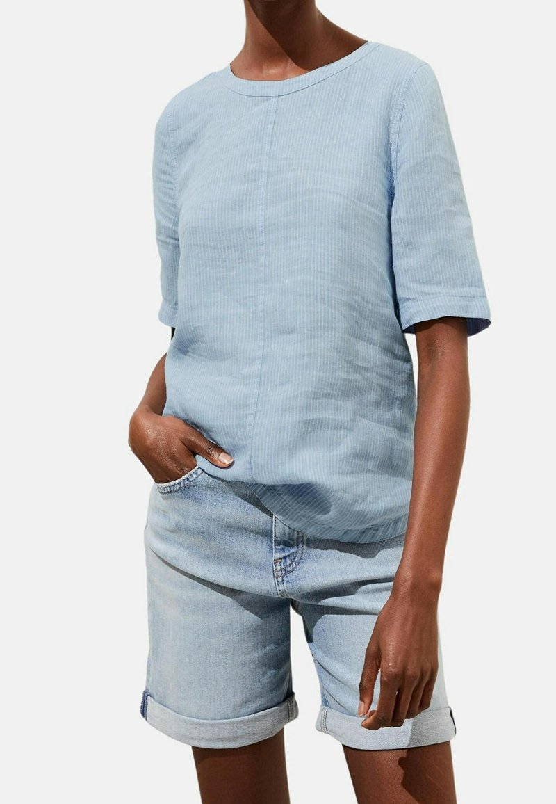 Personne portant une chemise texturée bleu clair à manches courtes et un short en denim clair retroussé, avec une main dans la poche.