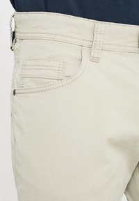 Pantalones cortos de algodón beige con una textura suave, que cuentan con un bolsillo frontal y costuras contrastantes a lo largo de los bordes. Incluye un cierre de botón metálico.