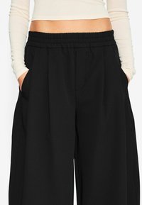 Schwarze weit geschnittene Stoffhosen mit elastischem Bund und seitlichen Taschen, die eine glatte Textur und eine lockere Passform bieten.