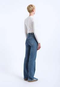 Blaue Jeans aus Denim mit geradem Schnitt, mittlerer Leibhöhe und Gesäßtaschen. Tragen mit einem eng anliegenden weißen Langarmshirt und blauen Sportschuhen.