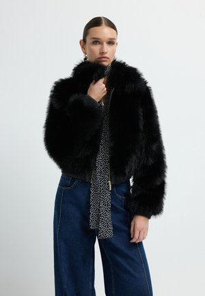 Black faux fur bomber jacket with a high collar and wide sleeves, featuring a zip closure and ribbed cuffs, worn over a polka-dot blouse.