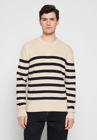 Maglione beige lavorato a maglia con righe blu navy, colletto a giro e polsini a costine. Presenta un piccolo logo sul petto. Indossato con pantaloni neri.