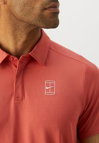 Rød polohefte laget av teksturert stoff, med en krage med to knapper og en hvit Nike-logo i et rektangulært ramme på venstre side.