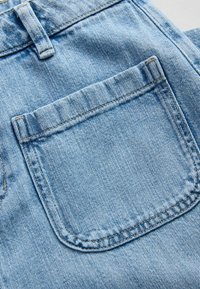 Közelkép világoskék farmeranyagból, amely hátsó zsebet és övhurkot ábrázol, látható varrással.