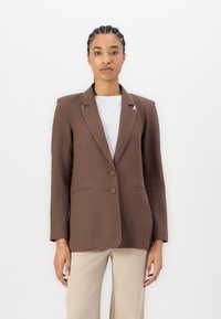 Ruske jakkesakko med en tettsittende silhuett, to knapper og sølvfargede detaljer på kragen. Kombinert med en hvit topp.